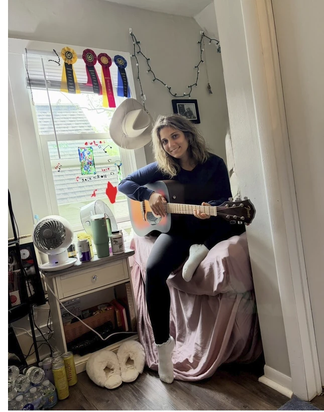 A smiling young adult seated with a guitar in her college room